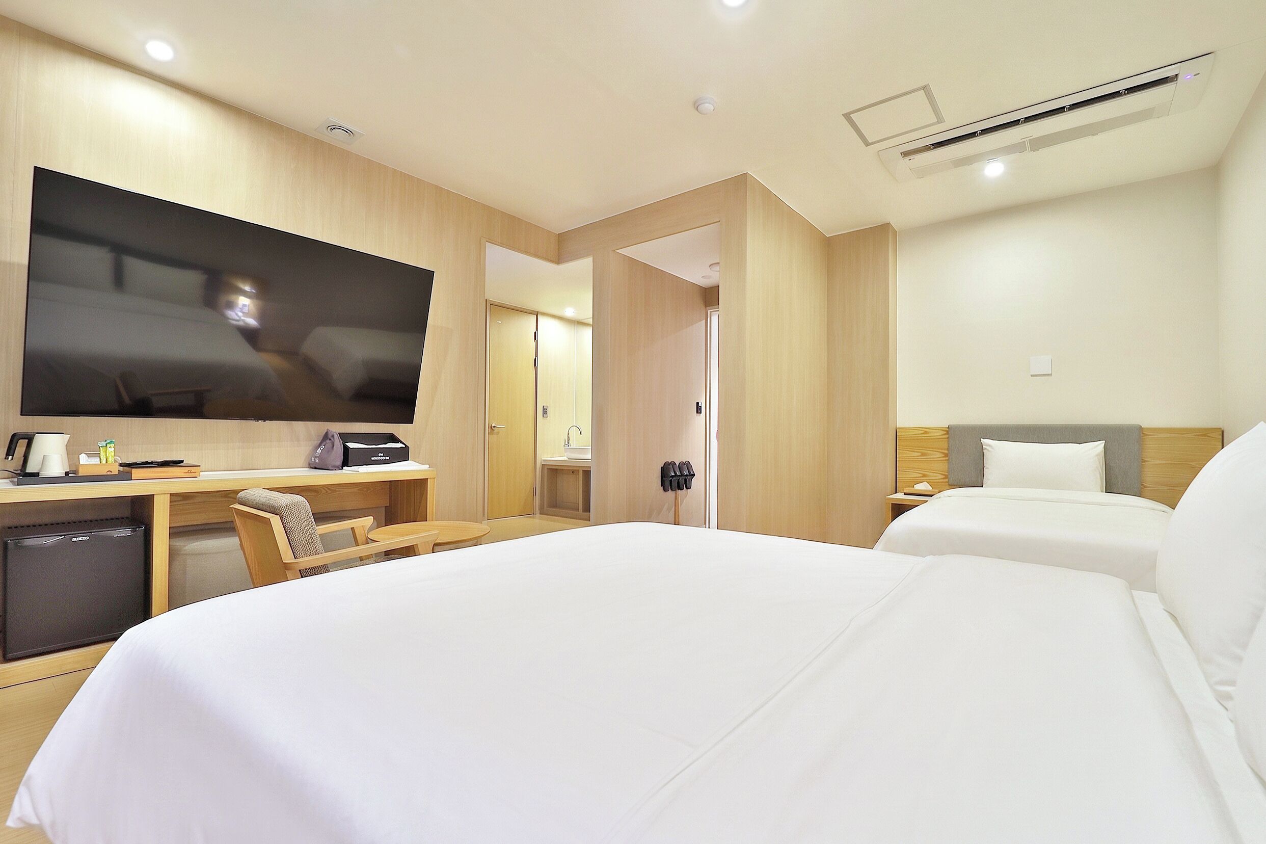 Habitación de hotel con cama, escritorio con televisor, refrigerador pequeño y baño con lavamanos y espejo.