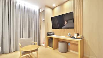 A hotel room with a wooden desk, a television mounted on the wall, a chair, a small ottoman, and a window with curtains.