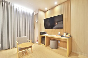 Habitación de hotel con un escritorio de madera, un televisor montado en la pared, una silla, un pequeño reposapiés y una ventana con cortinas.