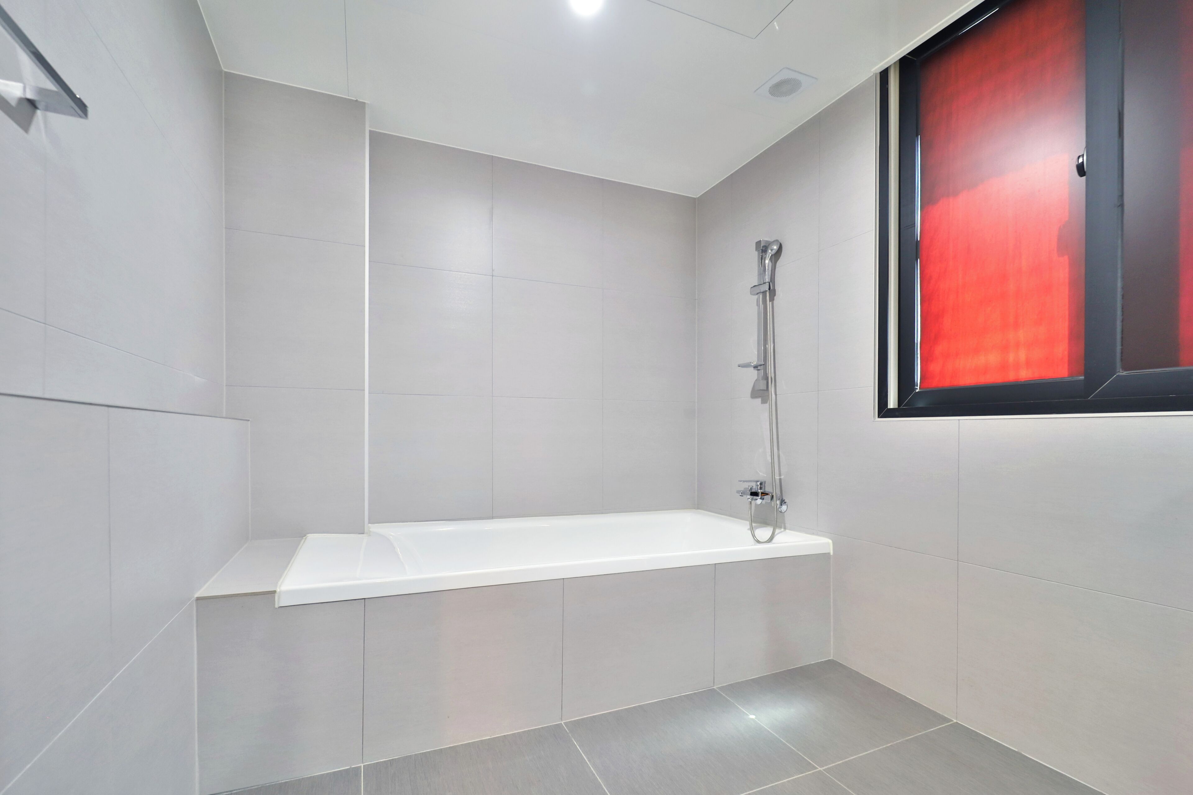 A modern bathroom with a white bathtub, a shower, and a red-framed window.