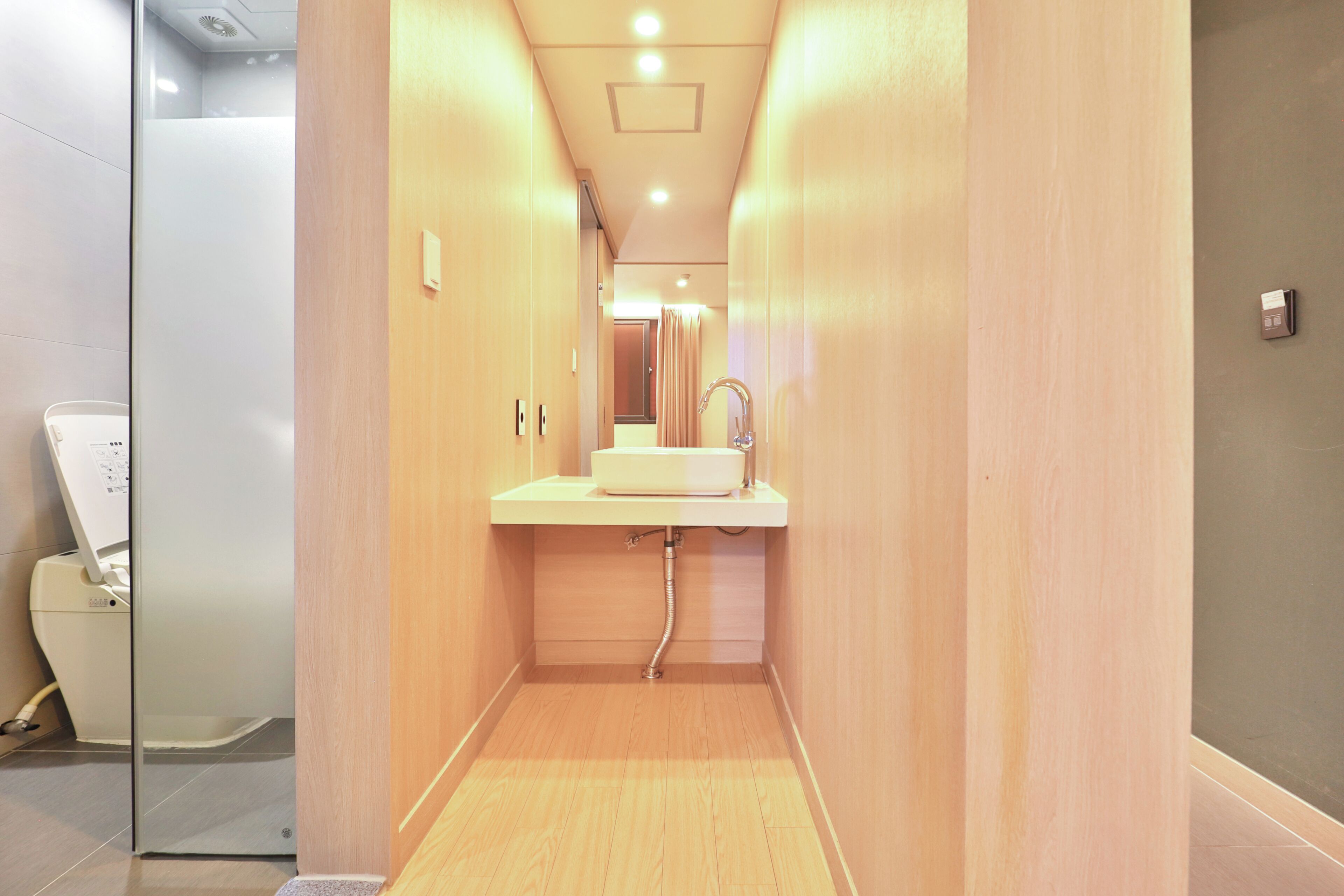 A narrow hallway with a wooden floor, a white sink, and a mirror on the wall.