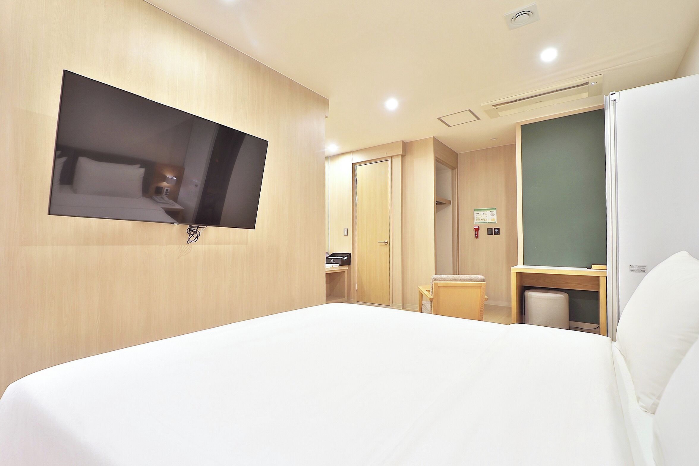 A hotel room with a large bed, a flat-screen TV mounted on the wall, a wooden wardrobe, a small desk with a chair, and a trash bin.