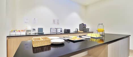 Un espace cuisine moderne avec un plan de travail noir, une machine à café, une bouilloire et un bol jaune posé sur un présentoir.