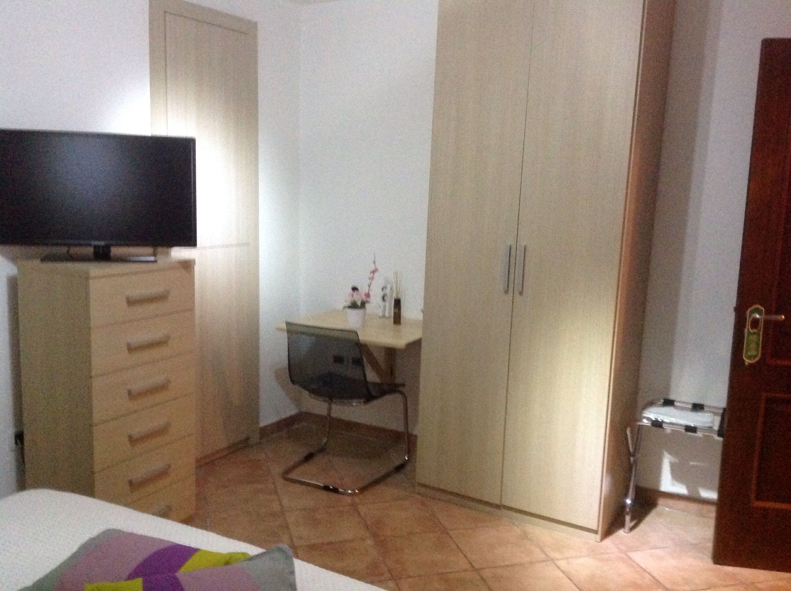 Un dormitorio con cama, cómoda, escritorio con silla, armario y televisor sobre la cómoda.