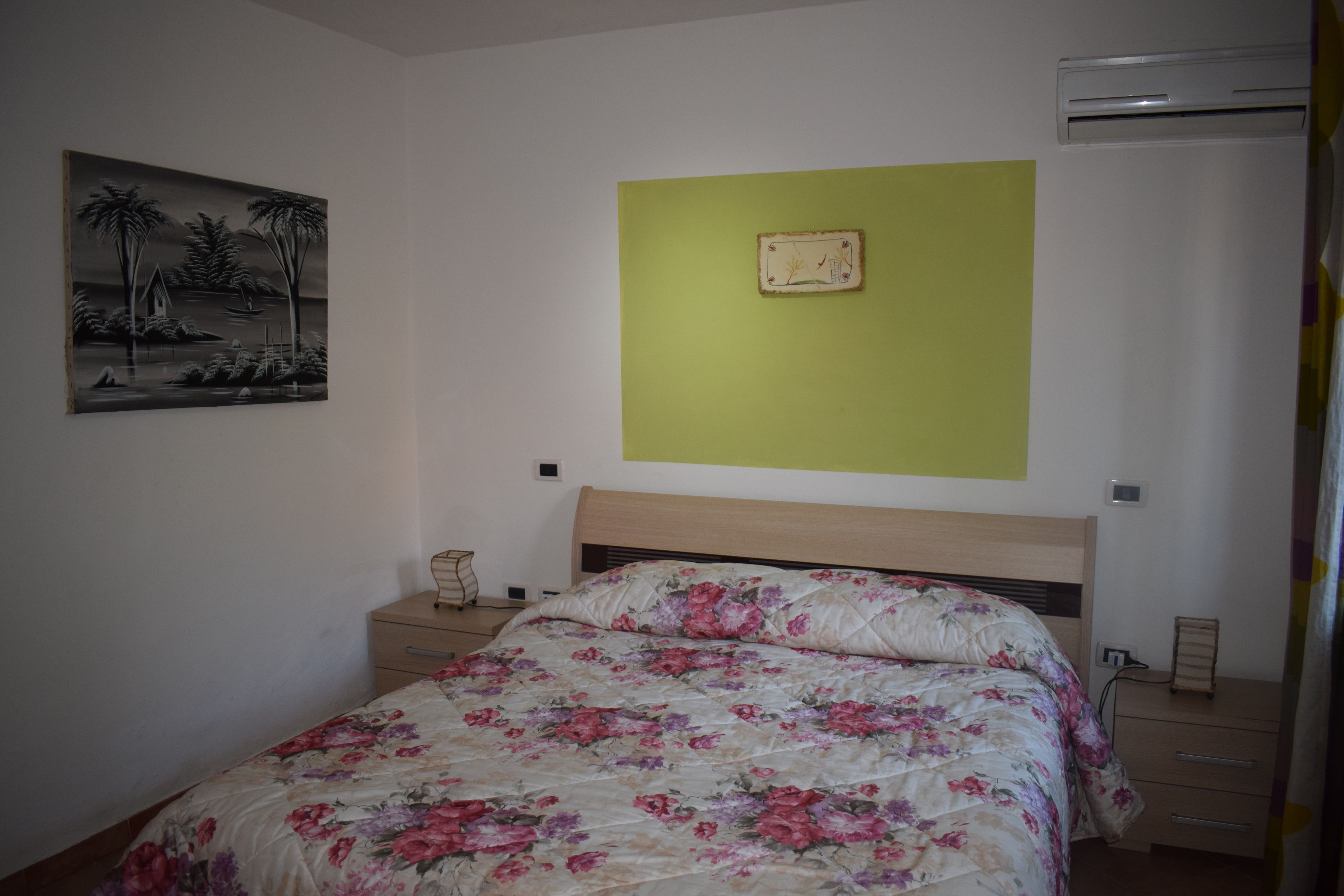 A bedroom with a bed, a nightstand, a wall-mounted air conditioner, and a framed picture.