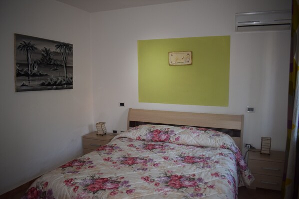 Un dormitorio con cama, mesita de noche, aire acondicionado instalado en la pared y un cuadro enmarcado.