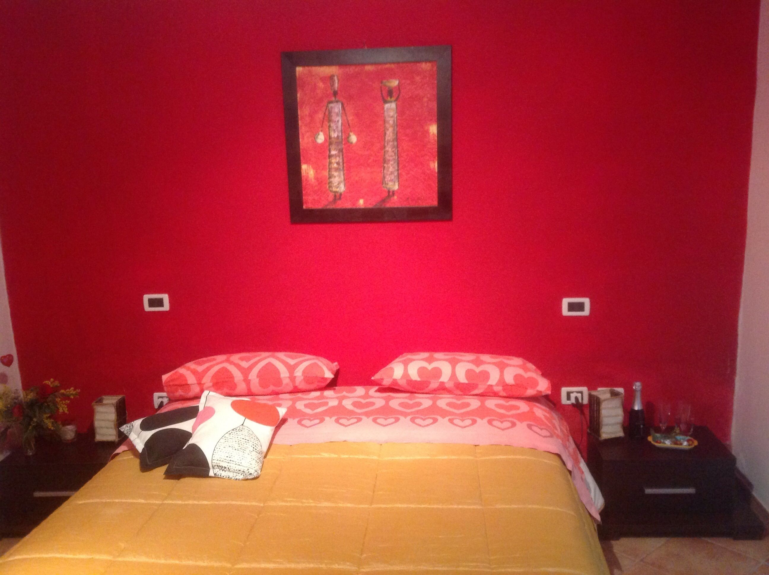 Un dormitorio con pared roja, una cama con almohadas estampadas, una obra de arte enmarcada sobre la cama y una mesita de noche con una botella y vasos.