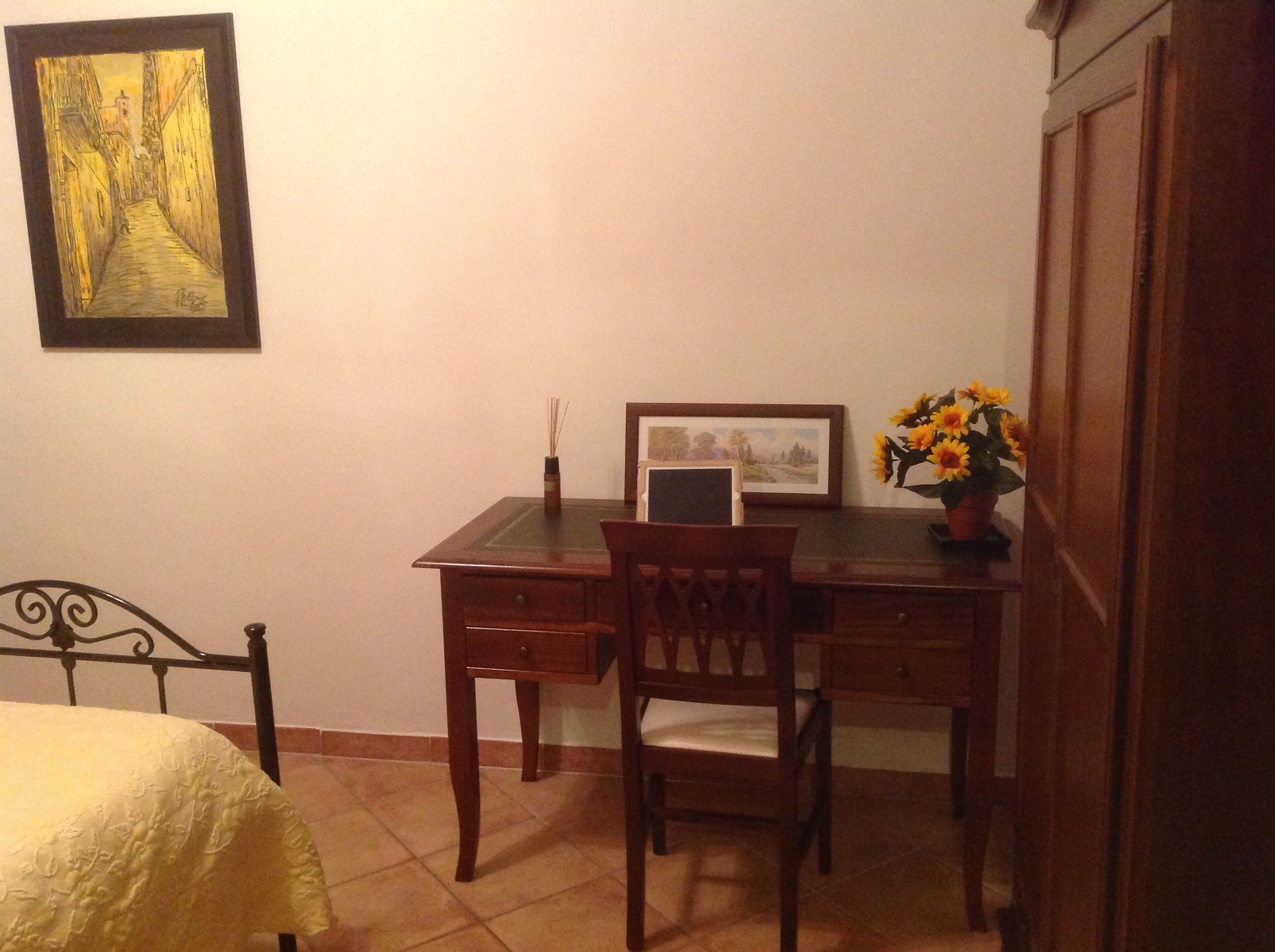 A room with a bed, a desk with a chair, a framed picture, a vase with flowers, and a painting on the wall.