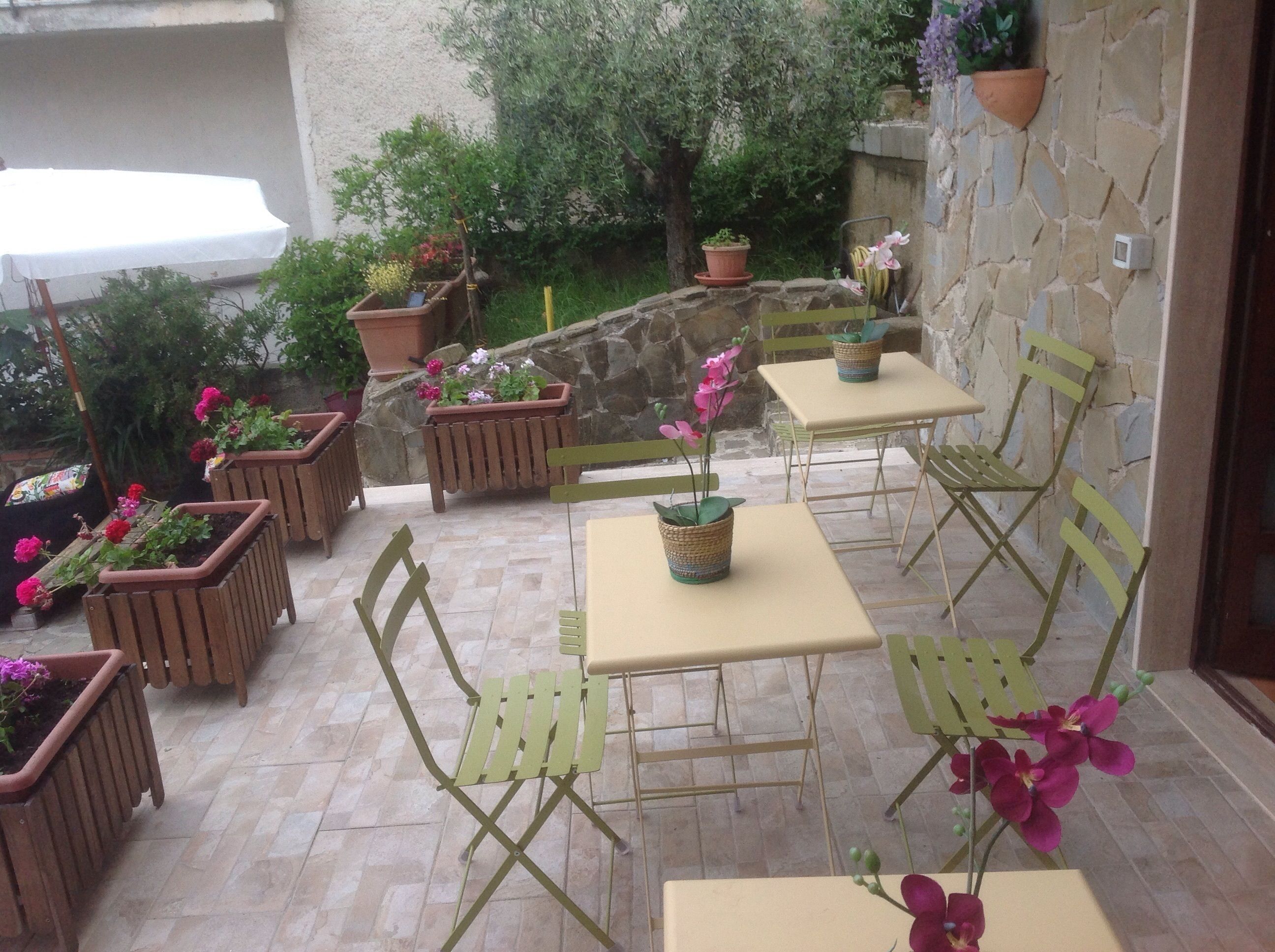 An outdoor patio with wooden chairs and tables, surrounded by potted plants and a stone wall.