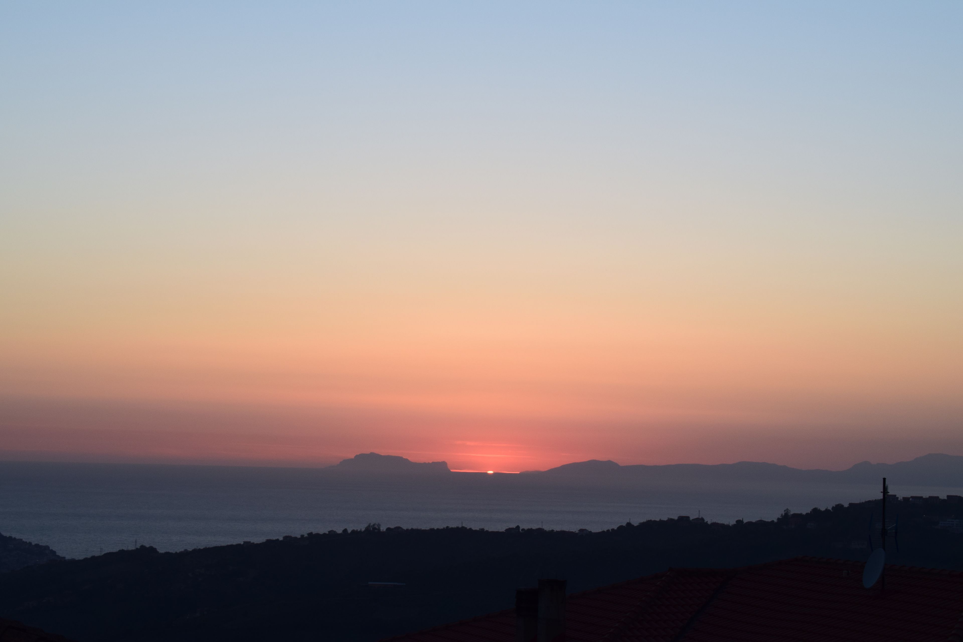 A sunset over a coastal landscape with a silhouette of hills and a satellite dish on a rooftop.