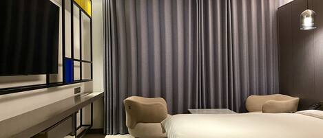 A modern hotel room with a large bed, a flat-screen TV mounted on the wall, a small table, two armchairs, and a large window with grey curtains.