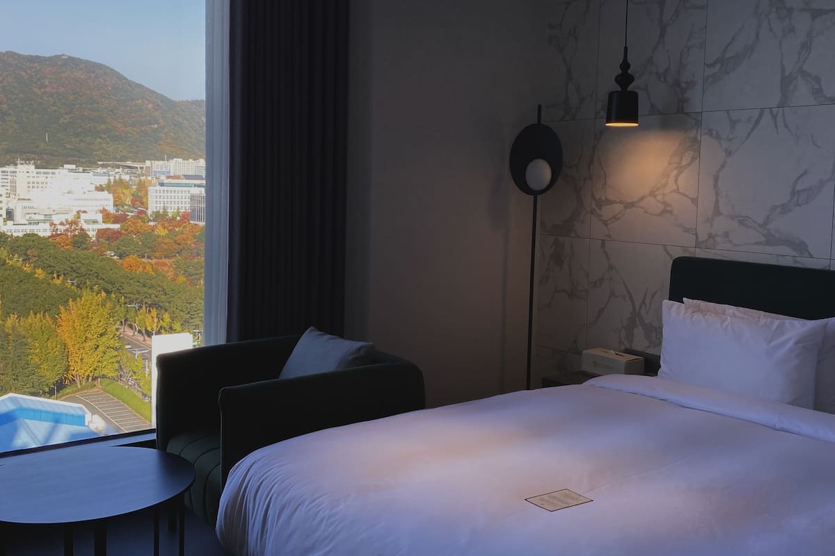 Habitación de hotel con cama, una silla, una mesa, una lámpara y vistas a un paisaje urbano y montañas.