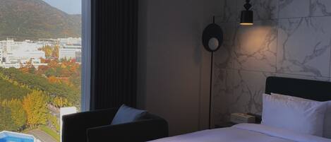 Habitación de hotel con cama, una silla, una mesa, una lámpara y vistas a un paisaje urbano y montañas.