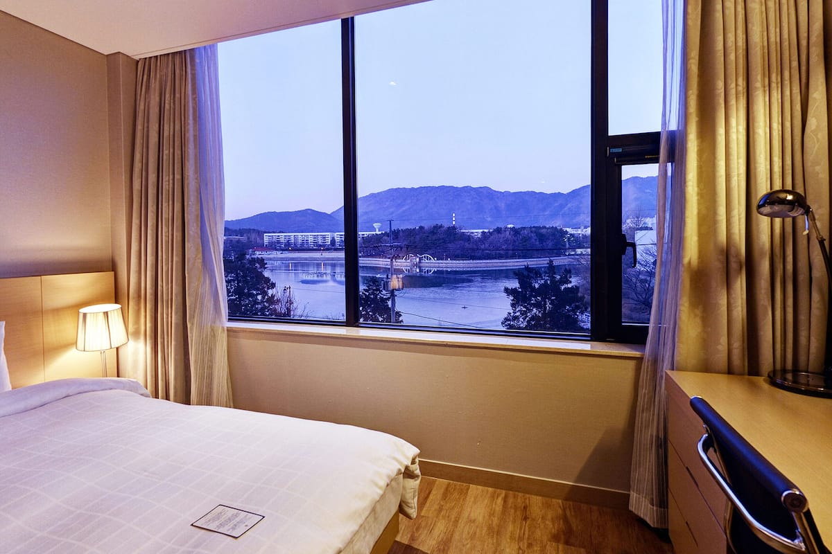 Una habitación de hotel con un ventanal grande, una cama, un escritorio y una lámpara.