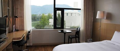 A hotel room with a bed, a desk with a chair, a TV, a window with a view of mountains, and a lamp.