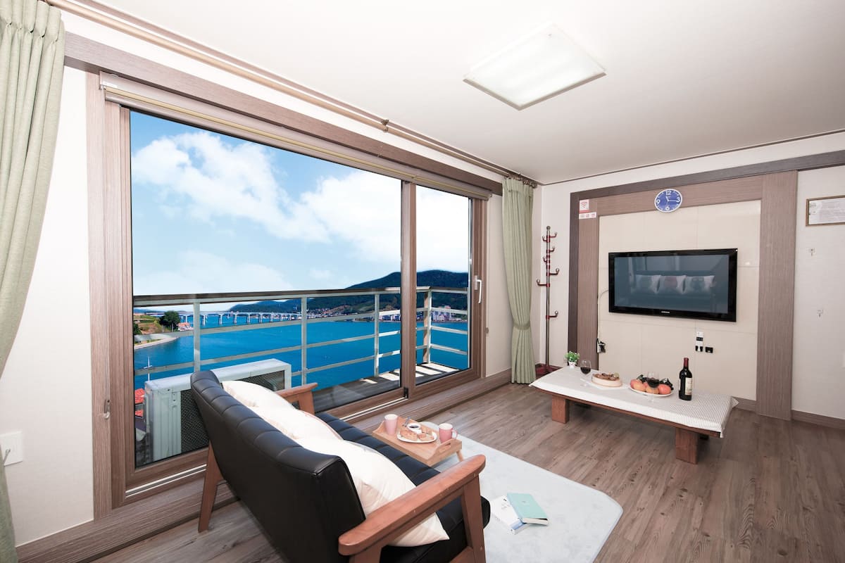 A balcony with a view of a body of water and mountains, a sofa, a table with food and drinks, and a TV mounted on the wall.