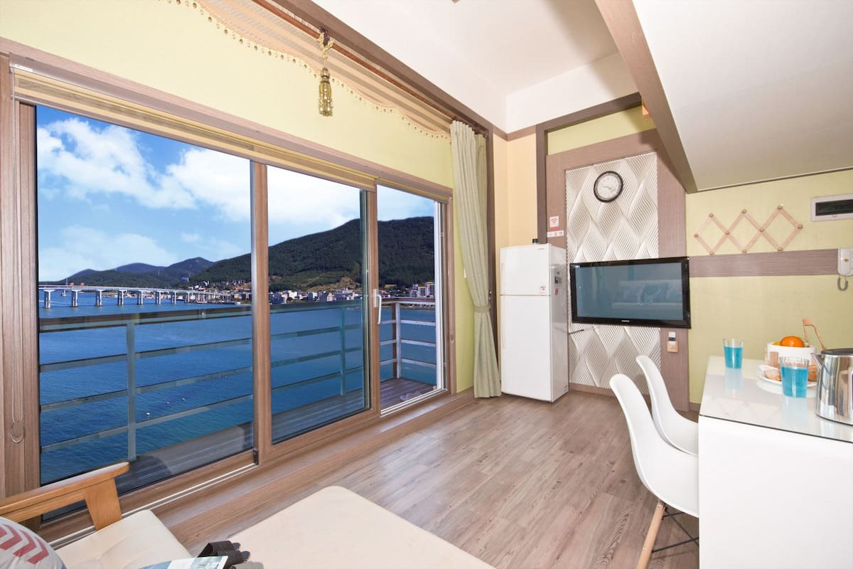 A room with a large sliding glass door leading to a balcony with a view of a bridge and water. There is a small refrigerator, a flat-screen TV mounted on the wall, and a dining table with chairs.