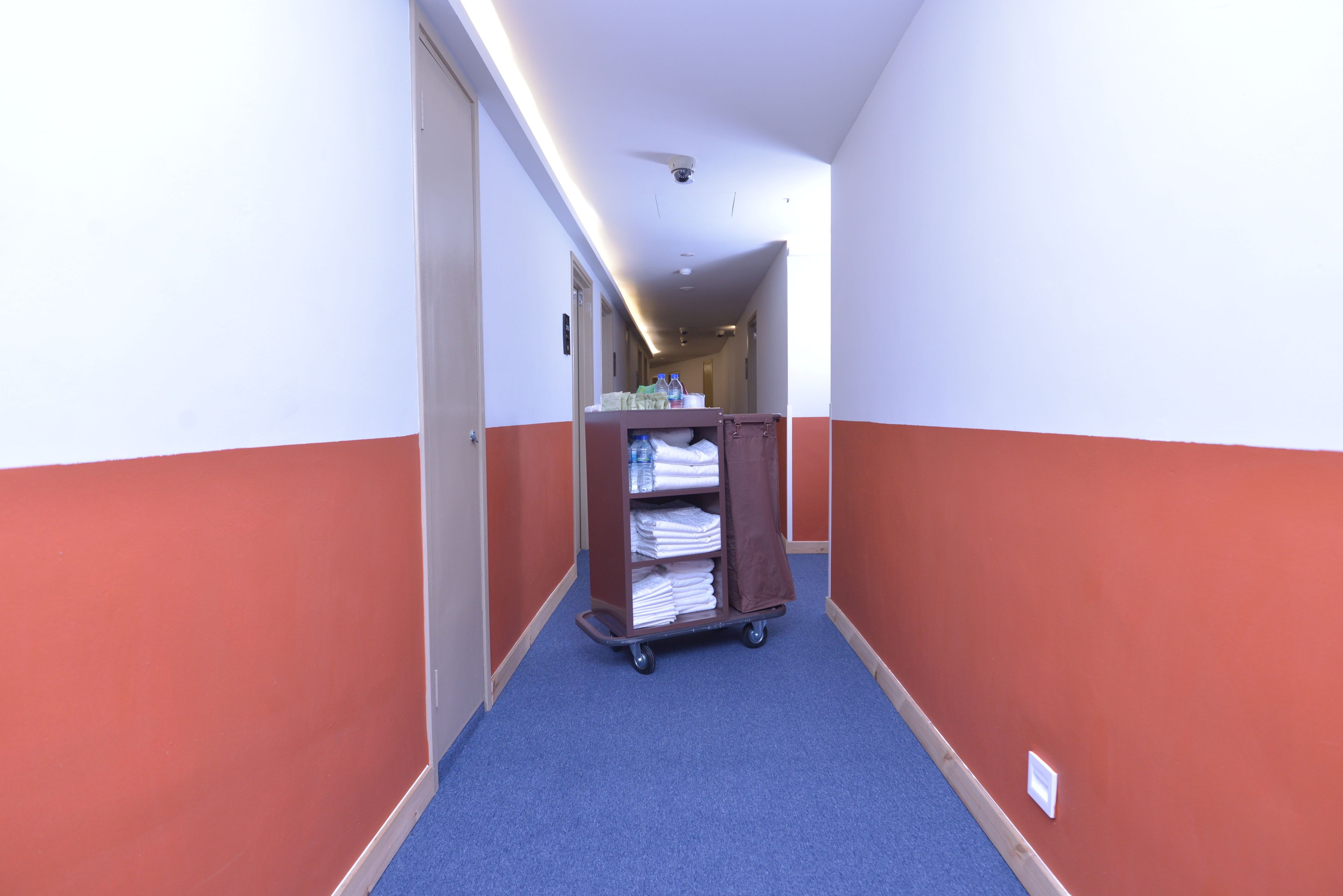 Un couloir d’hôtel avec un tapis bleu, un chariot marron contenant des serviettes pliées et un mur rouge et blanc.