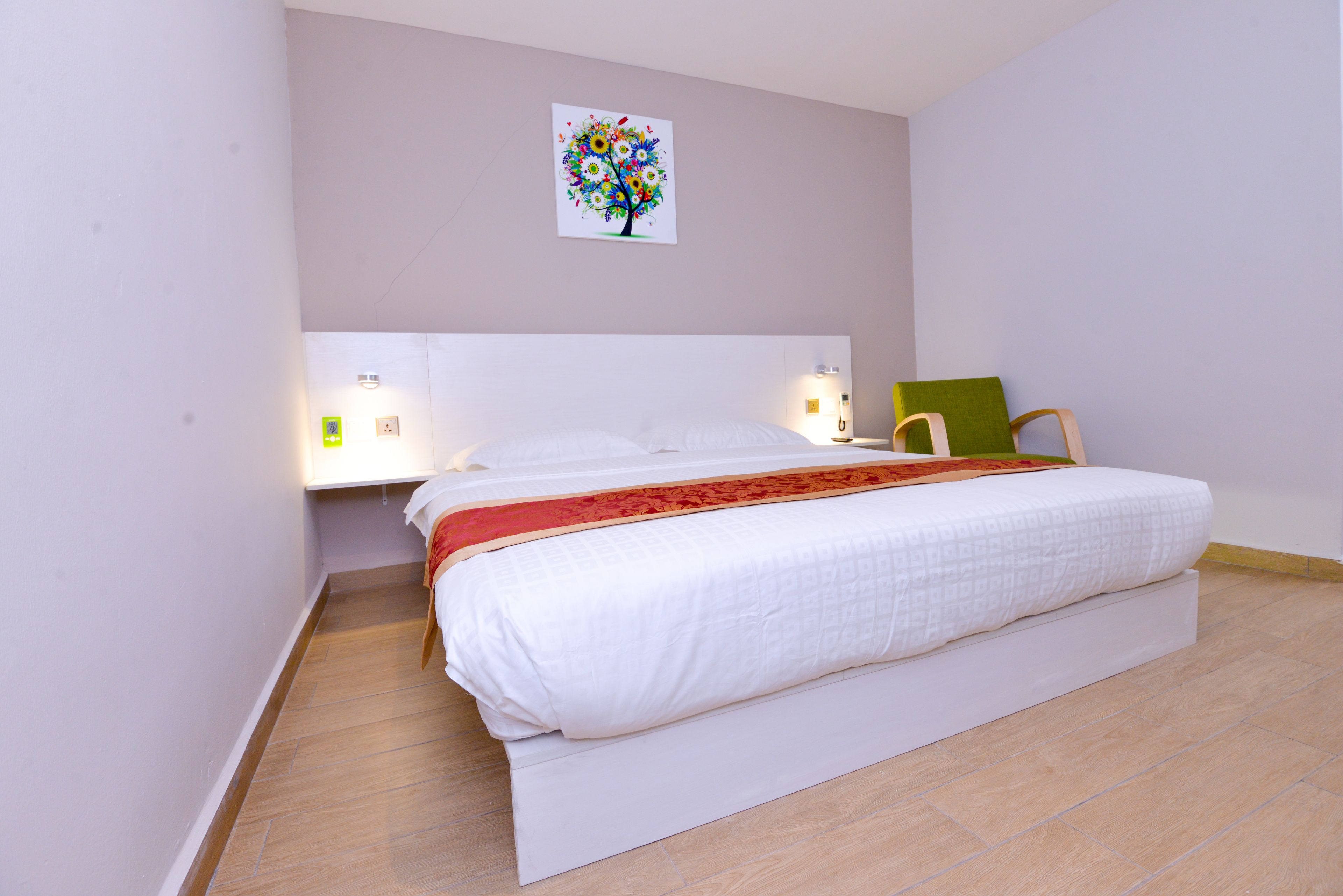 A neatly made bed with a white comforter and a red blanket, a green pillow, and a wall-mounted artwork of a tree.
