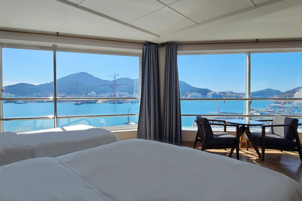 Habitación de hotel con ventanas amplias que ofrecen vista a un puerto, barcos y montañas distantes.
