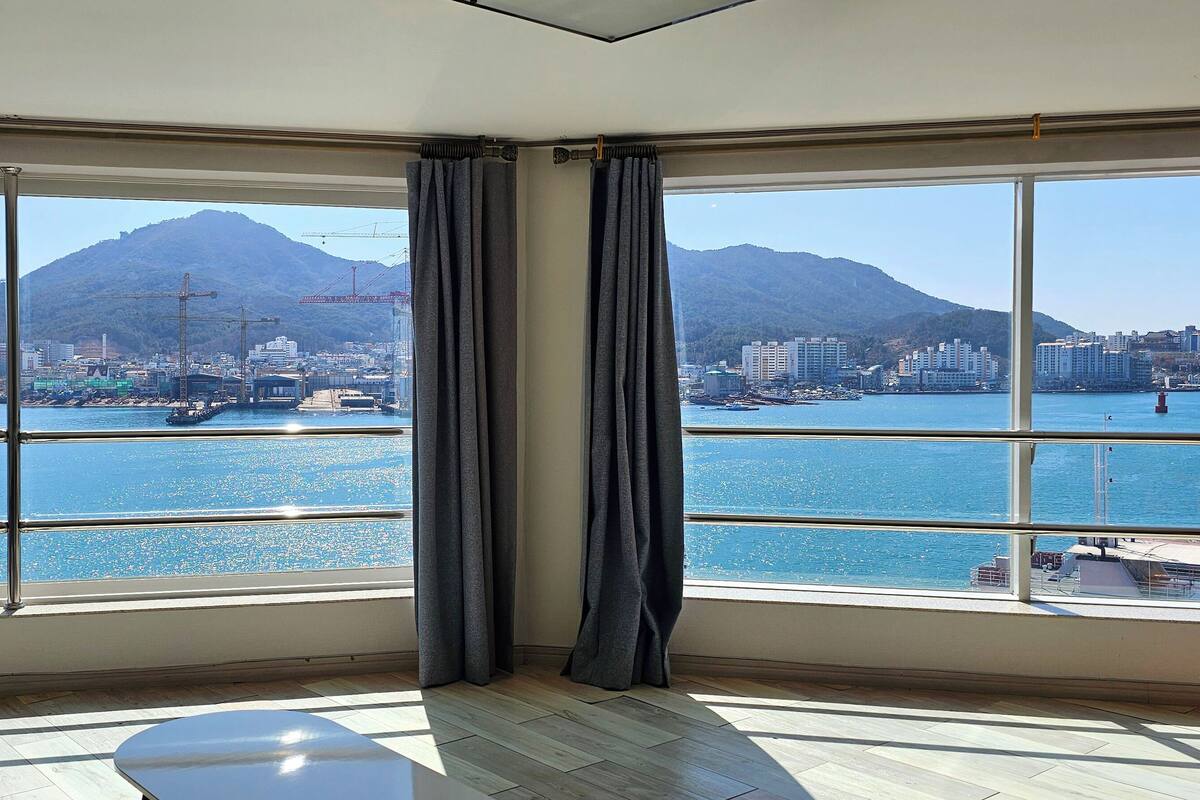 Una habitación con grandes ventanales que ofrecen vistas a un puerto, barcos y un paisaje montañoso.