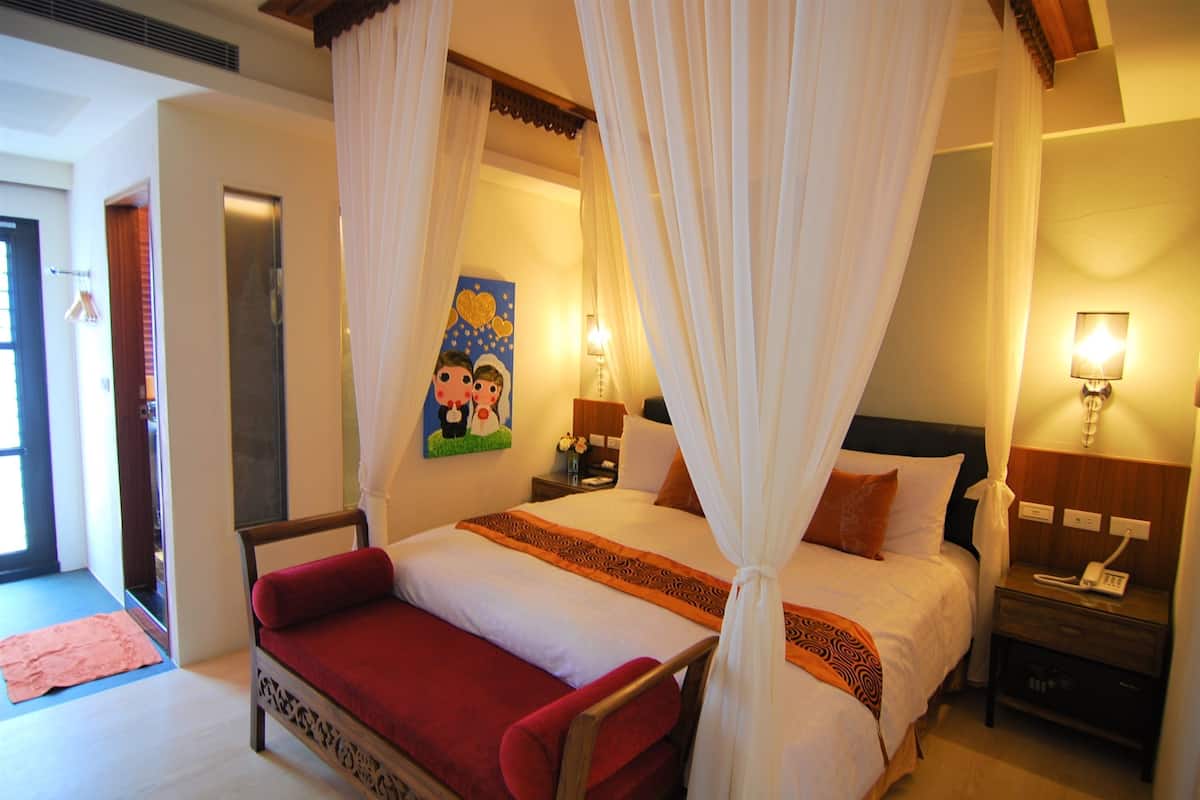 A hotel room with a bed, a nightstand with a lamp, a painting on the wall, and a door.