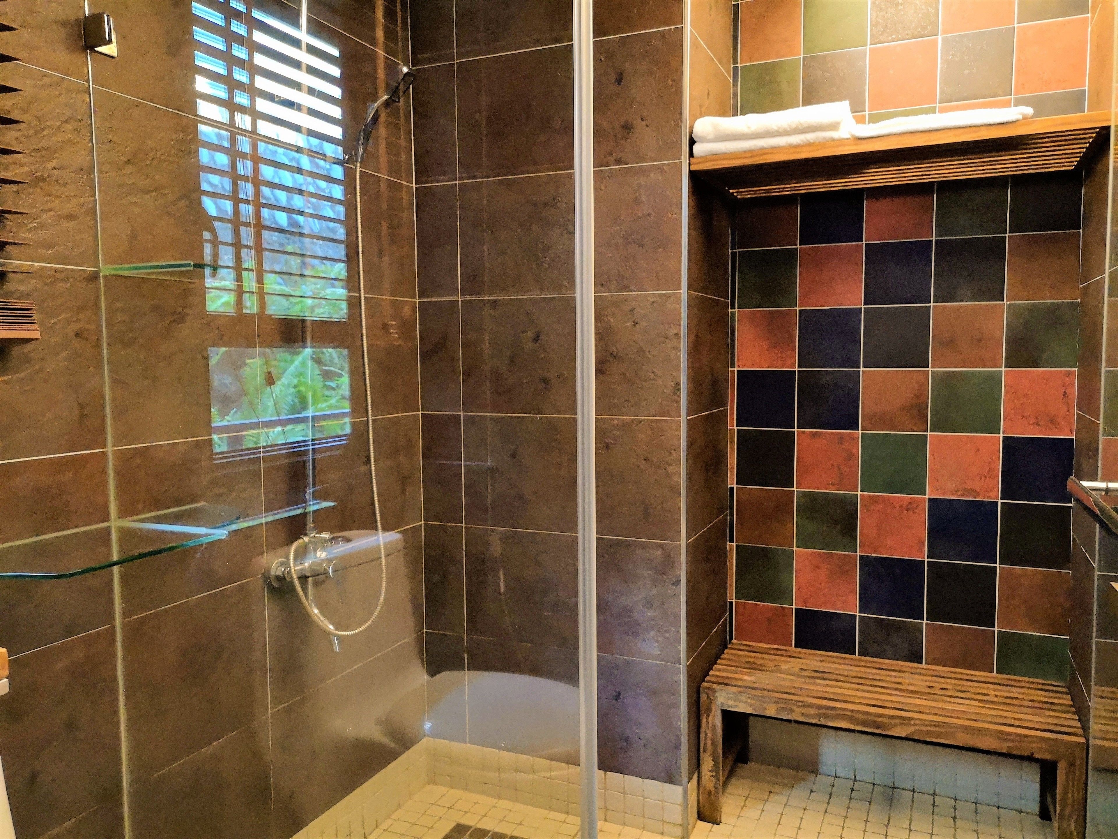 Un área de ducha con mampara de vidrio, un banco de madera y un muro con baldosas de diseño de cuadros.