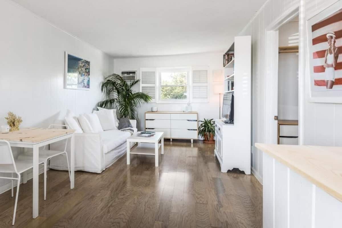 A modern, white-themed living room with a wooden floor, a white sofa, a small table, and a kitchen area with white cabinets and a window.