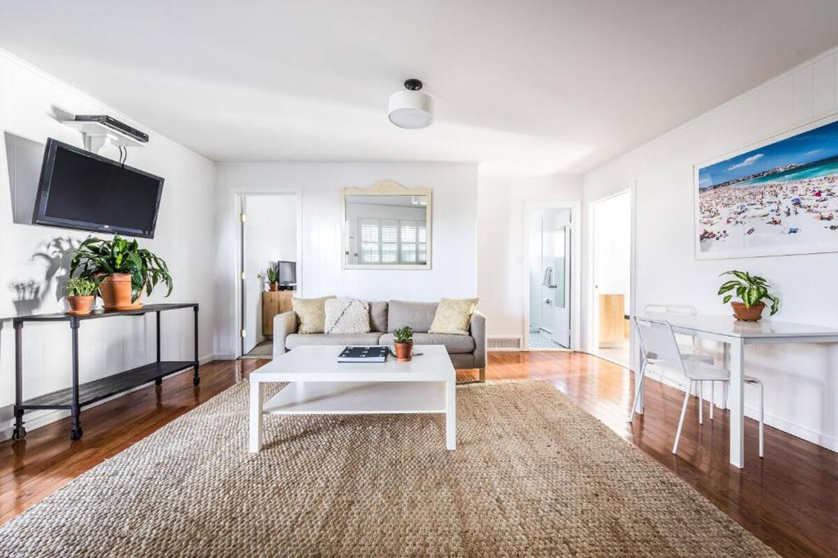 A modern living room with a white sofa, a coffee table, and a TV mounted on the wall. There are potted plants and a painting on the walls.