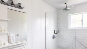 Une salle de bain moderne avec une douche à l’italienne en verre, des murs recouverts de carreaux blancs, un grand miroir et une fenêtre.