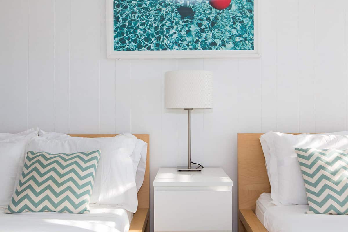 A hotel room with two beds, each with white bedding and a green and white chevron pillow. A white nightstand with a lamp and a framed picture of a swimming pool is between the beds.