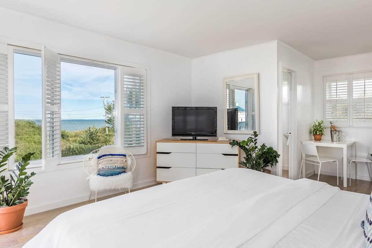 A bedroom with a bed, a desk, a chair, a TV, and a view of the sea.
