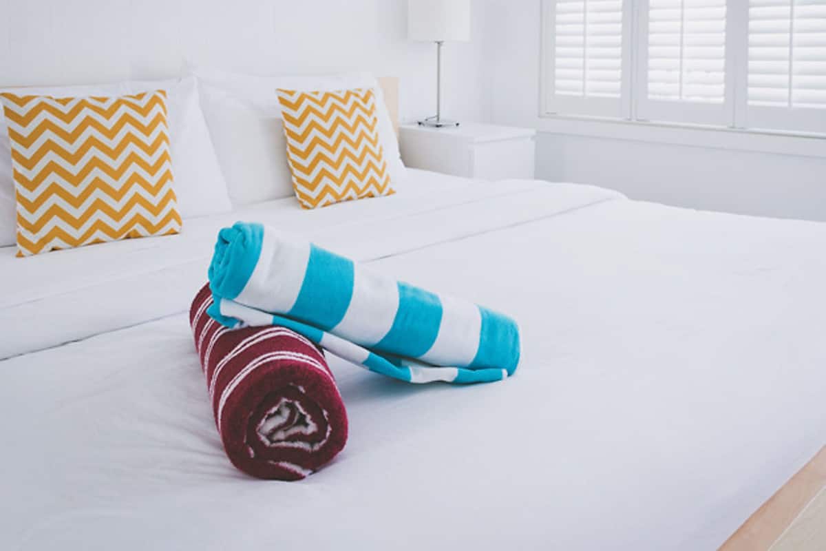A neatly made bed with white bedding, two yellow and white chevron pillows, and a rolled towel in blue and white stripes.