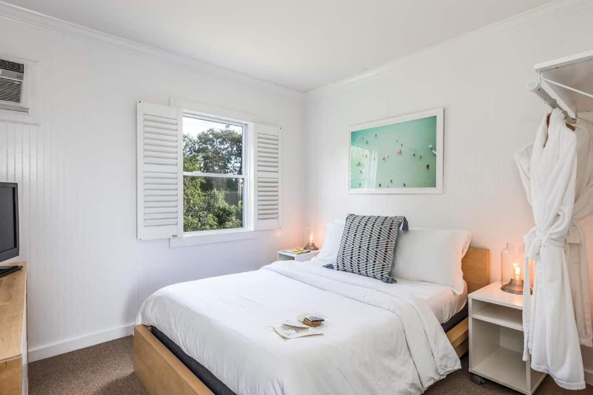 A bedroom with a bed, a nightstand, a TV, a window with shutters, a painting, and a robe hanging on a rack.
