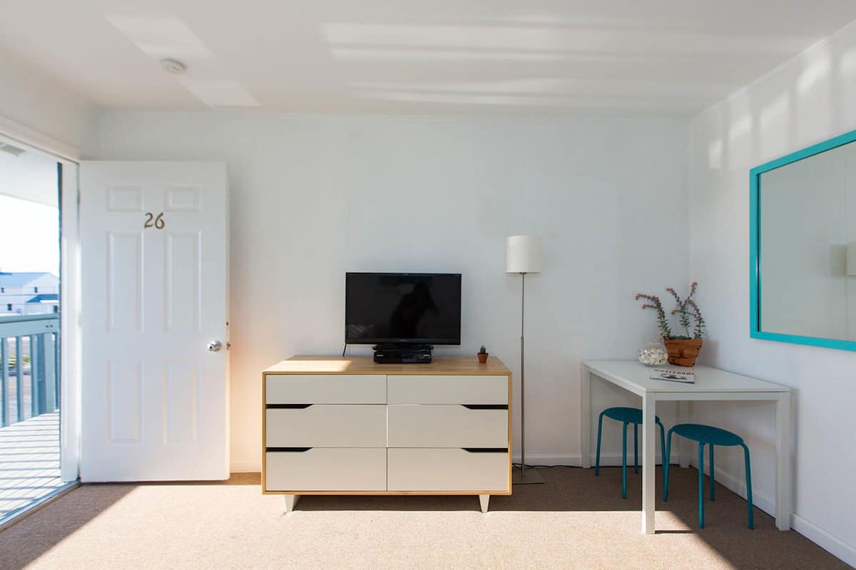 A modern room with a white door marked "26", a television on a wooden cabinet, a desk with two chairs, and a large mirror on the wall.