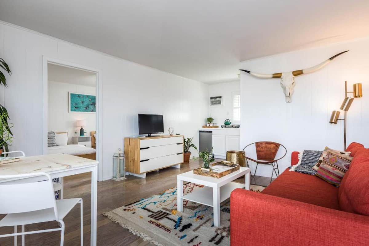 A modern living room with a red sofa, white coffee table, and a TV. There's a kitchen in the background with white cabinets and a dining area with a white table and chairs.