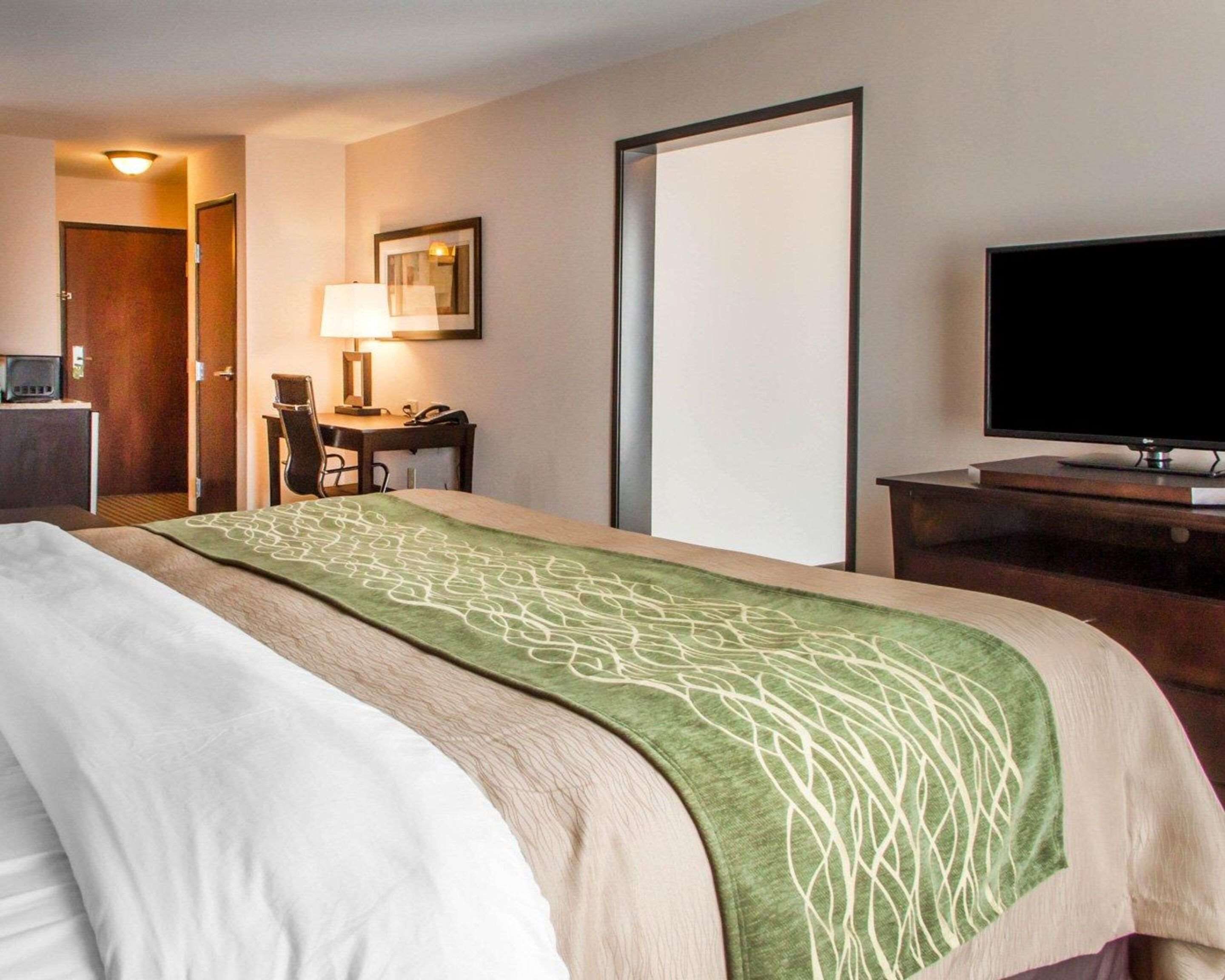 Quarto de hotel com uma cama, uma escrivaninha, uma cadeira, uma televisão e uma porta.