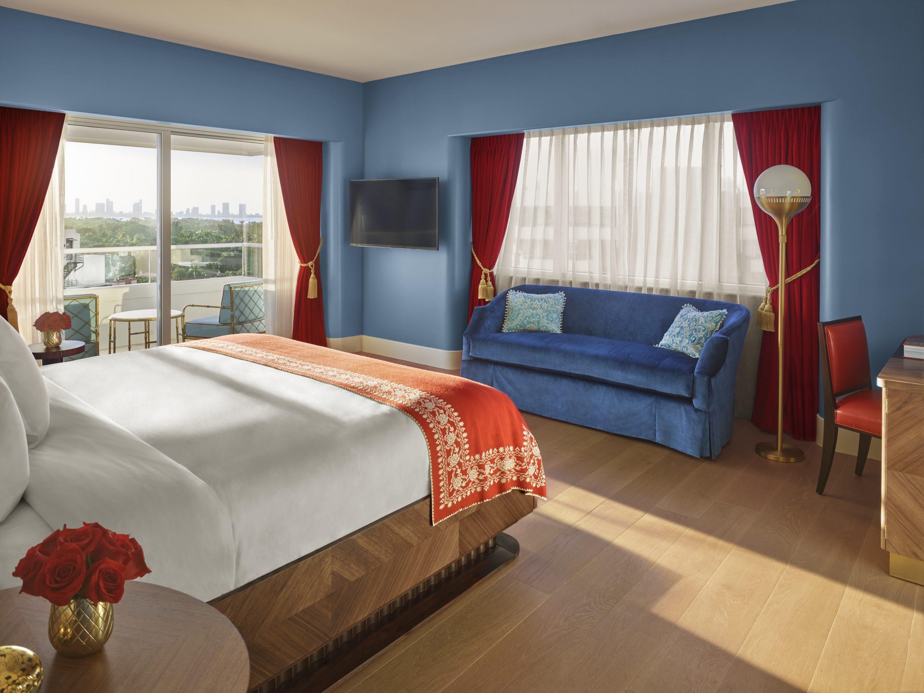 Habitación de hotel con cama, sofá, televisor, balcón con vistas y lámpara de pie.