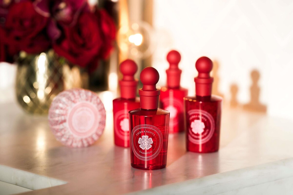 A set of red shampoo bottles with a logo, placed on a marble surface with a blurred background of red roses and a vase.