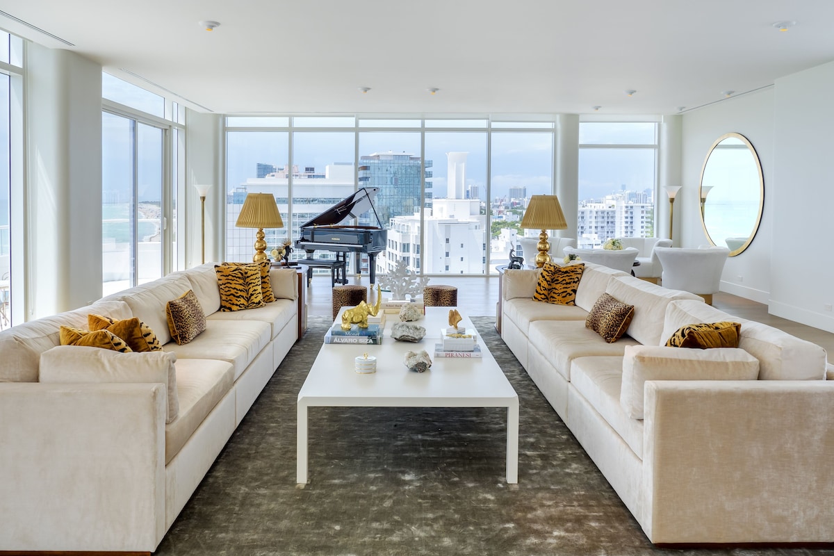 A spacious living room with two white sofas, a central coffee table, and a grand piano. Large windows offer a view of the city and the ocean.