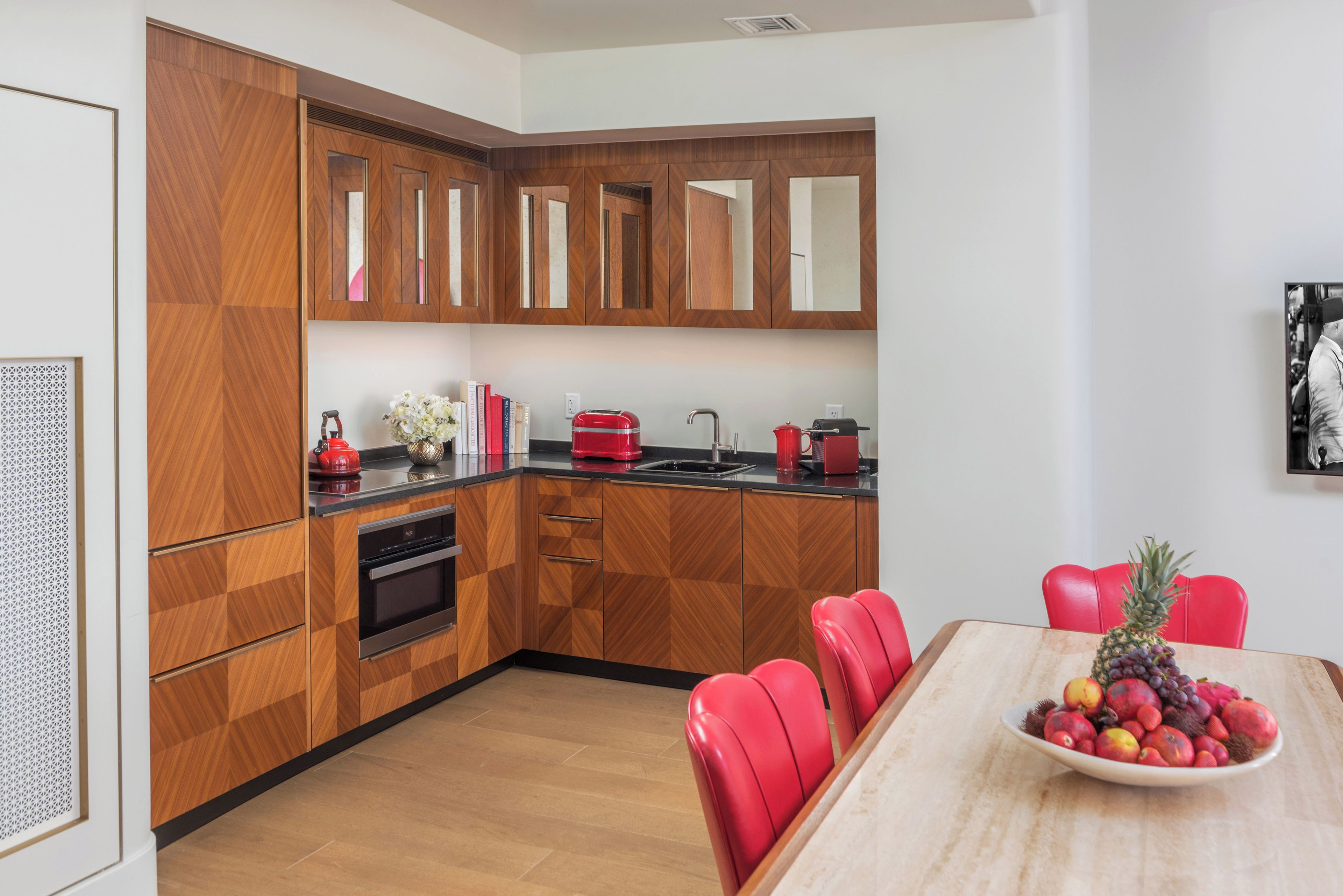 Una cocina moderna con armarios de madera, horno empotrado y comedor con una mesa y sillas rojas.