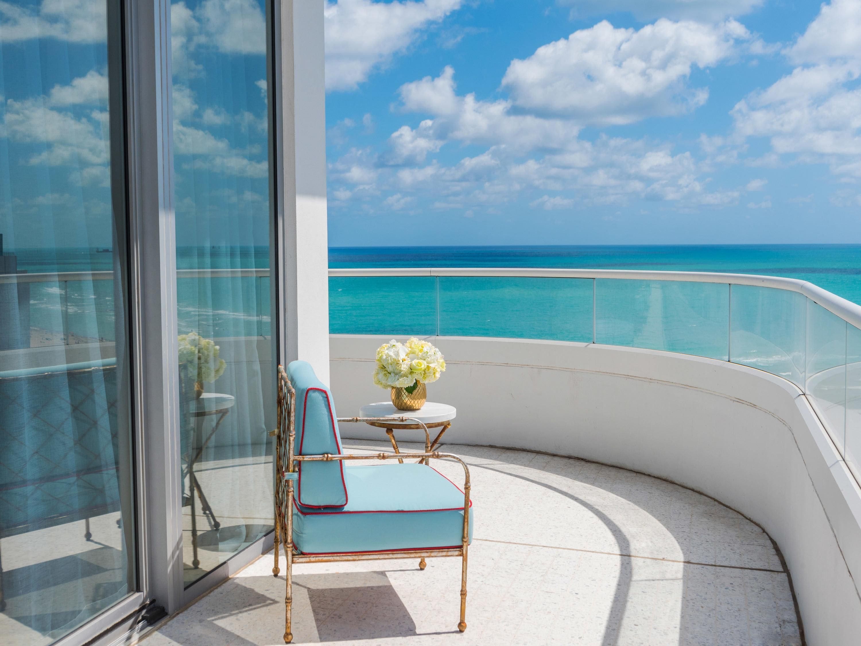 A balcony with a view of the ocean, a chair, a small table, and a vase of flowers.