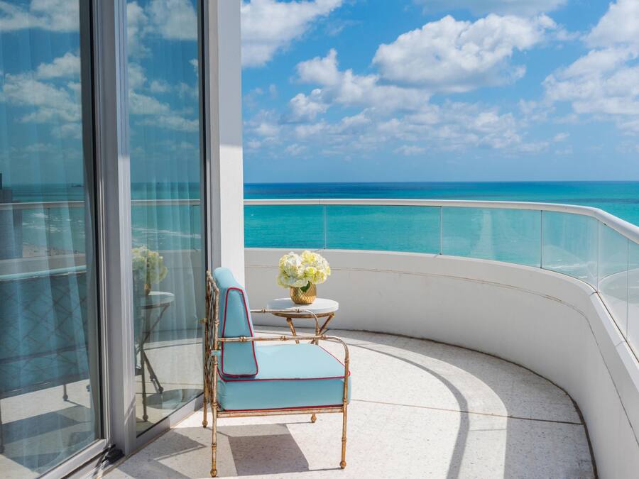 A balcony with a view of the ocean, a chair, a small table, and a vase of flowers.