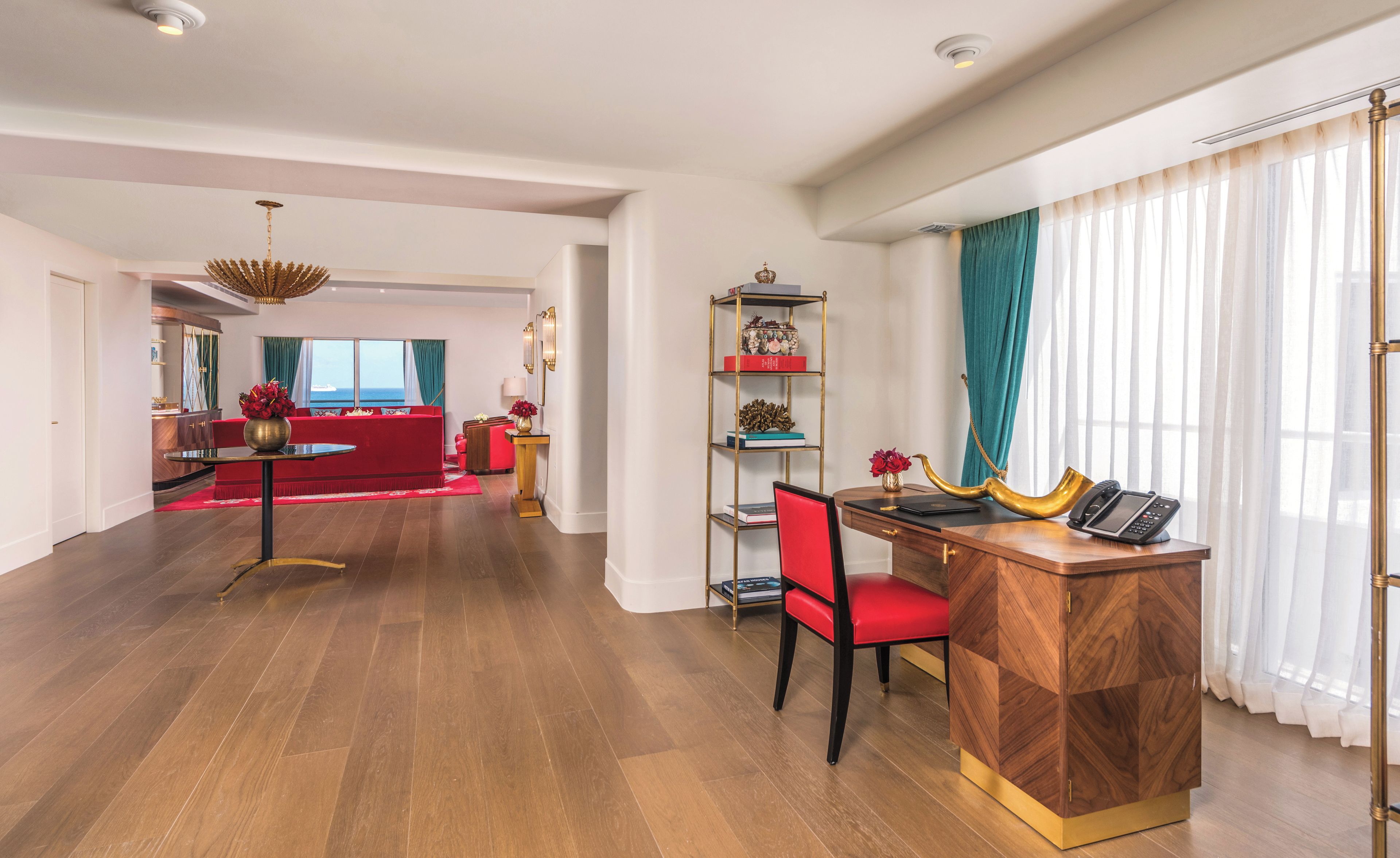 Un amplio espacio de sala con un sofá rojo, una mesa de centro de madera y un escritorio con una silla. Hay una estantería con objetos decorativos y un ventanal con cortinas.