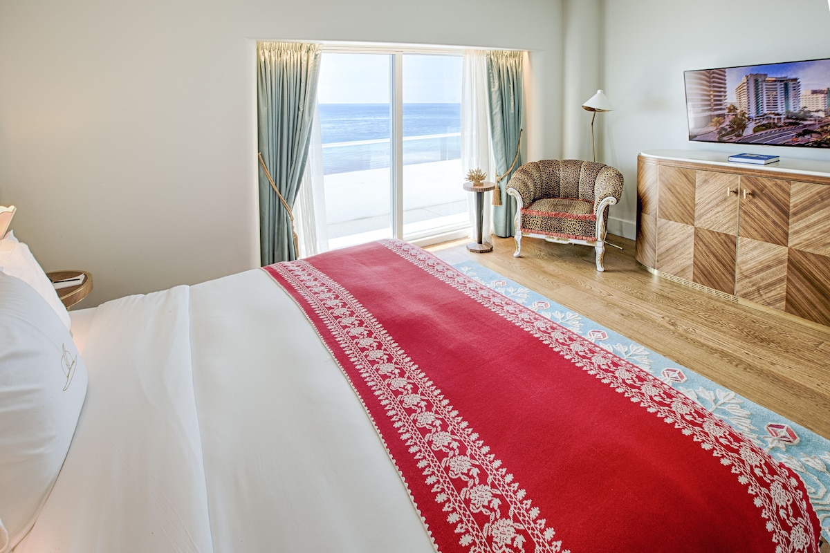A hotel room with a large bed, a TV, a chair, and a view of the ocean.