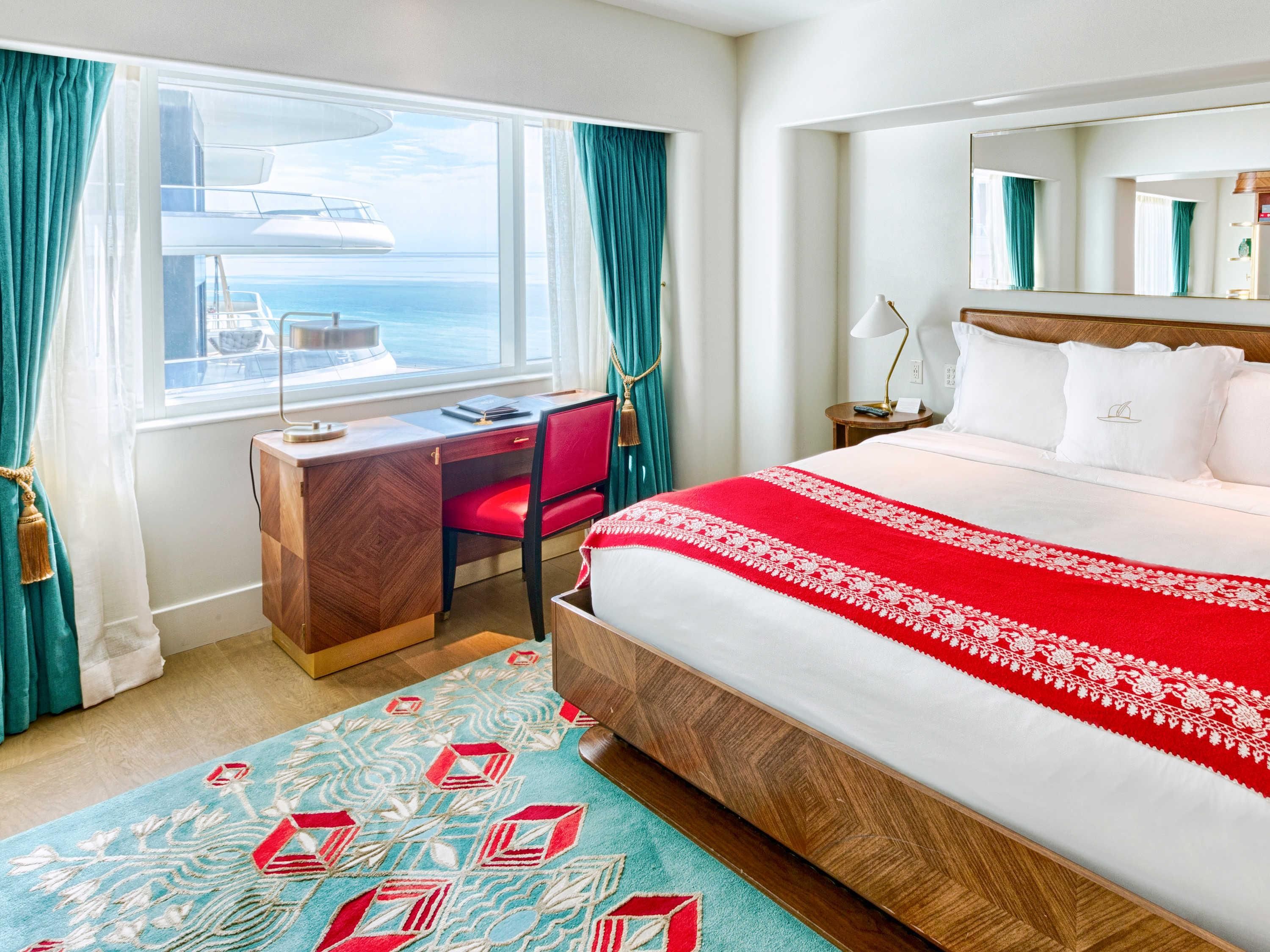 A hotel room with a large bed, a desk, a chair, a window with curtains, a nightstand, and a mirror.