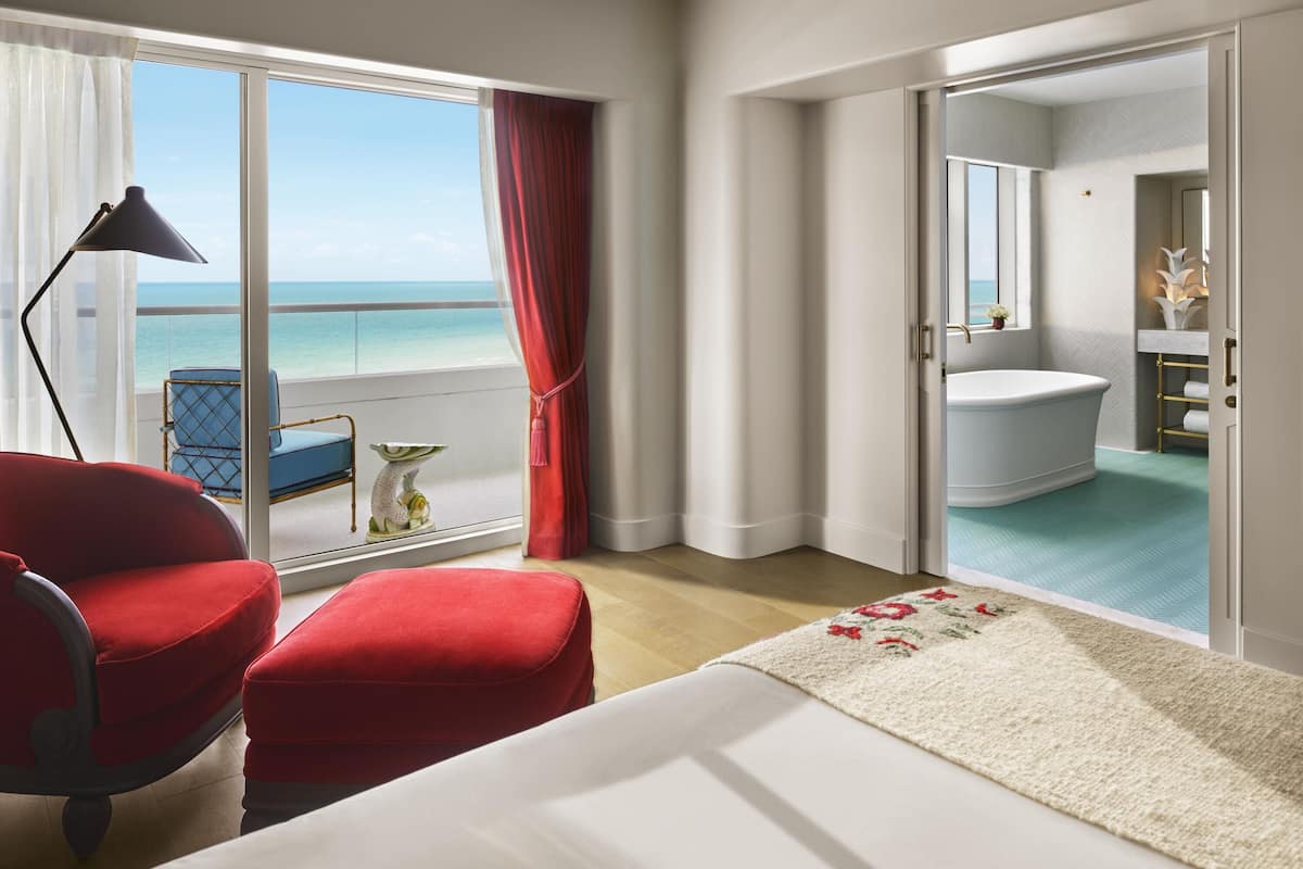 Ett hotellrum med en stor säng, en röd fåtölj, ett skrivbord och utsikt över havet.