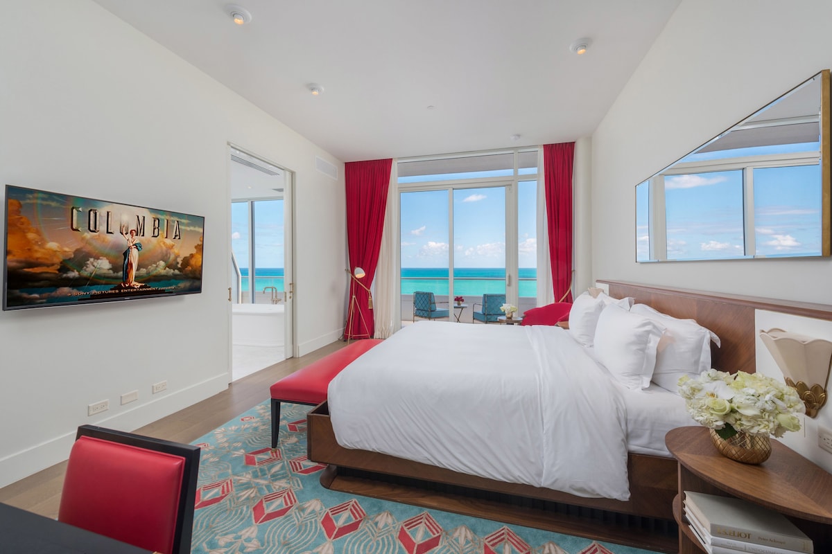 A hotel room with a large bed, a TV mounted on the wall, a view of the sea, and a red curtain.