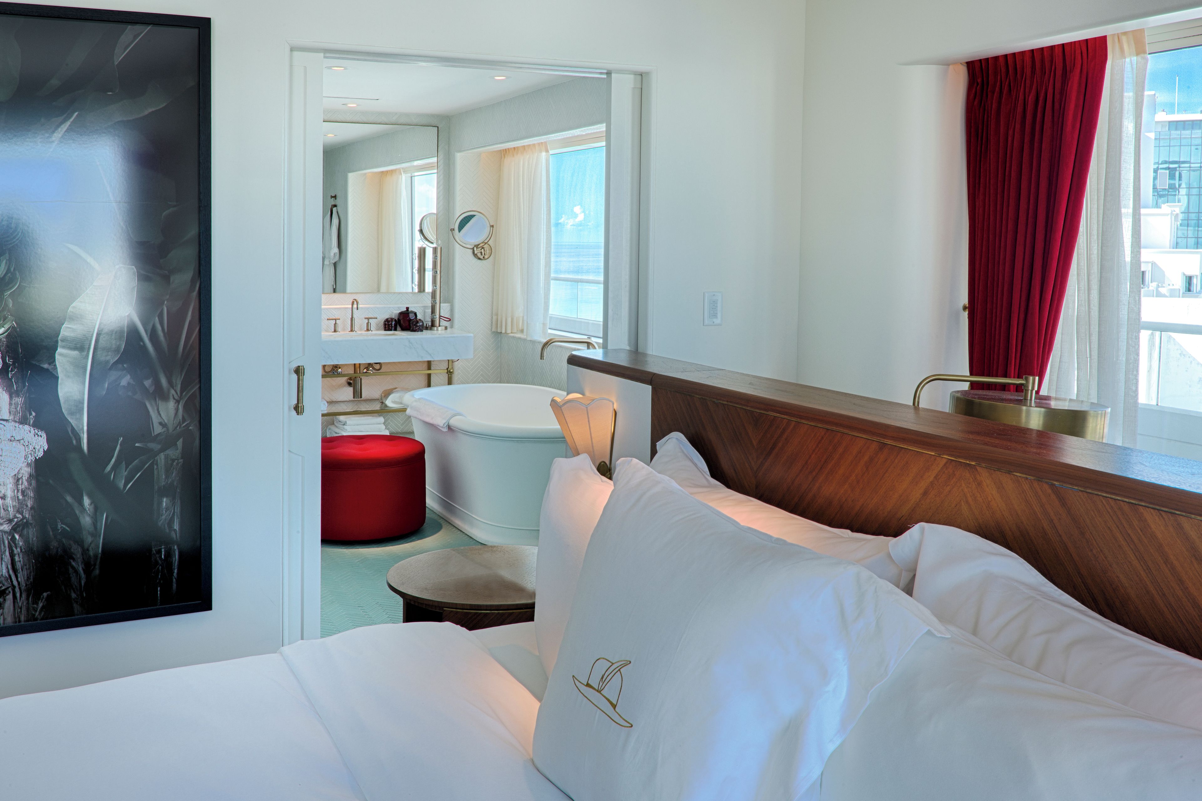 A hotel room with a bed, a red ottoman, a bathroom with a bathtub, and a large window with red curtains.