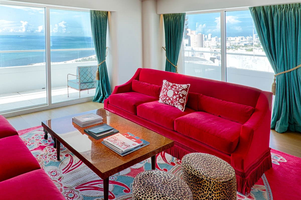 A red sofa with a patterned pillow, a glass coffee table with books, and a view of the ocean and cityscape through large windows with teal curtains.